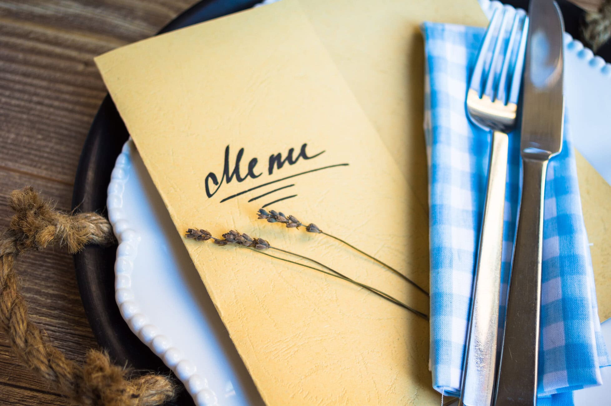 Close-up of a yellow menu card with the word 'menu' beside a fork and knife on a plate with a blue napkin, rustic table setting.