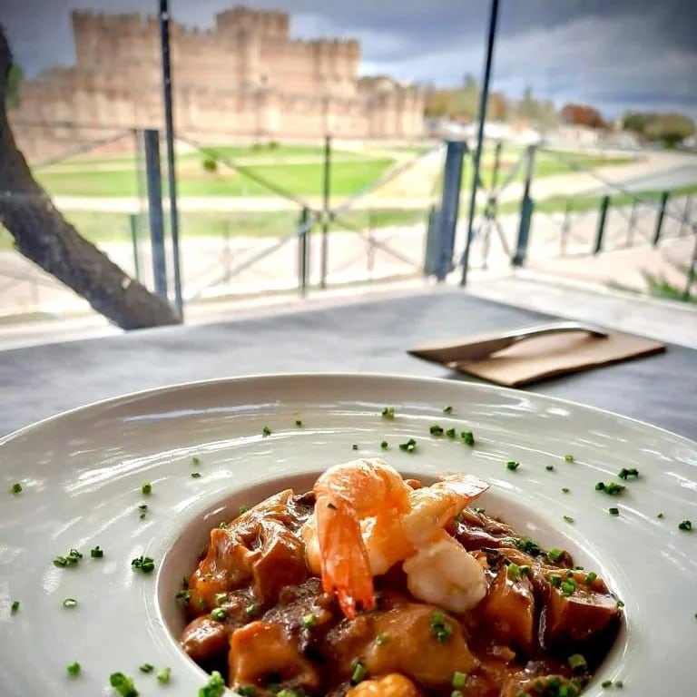 Shrimp and mushroom dish in a brown sauce, topped with a shrimp and chives, served on a white plate outdoors near a castle-like building