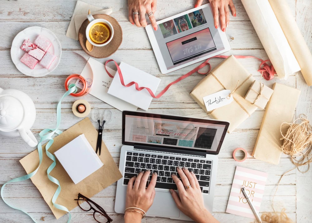 Overhead view of a laptop and tablet among wrapped gifts, ribbons, and a coffee on a bright desk, suggesting online holiday gifting.