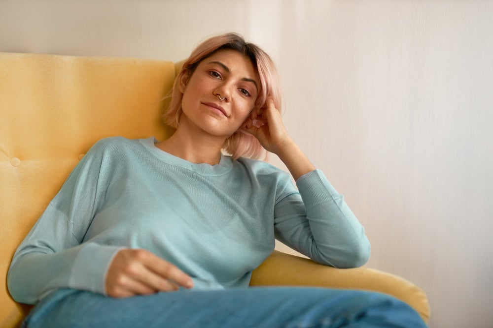 Person with pink hair and nose ring lounging on a yellow sofa in a blue sweater.