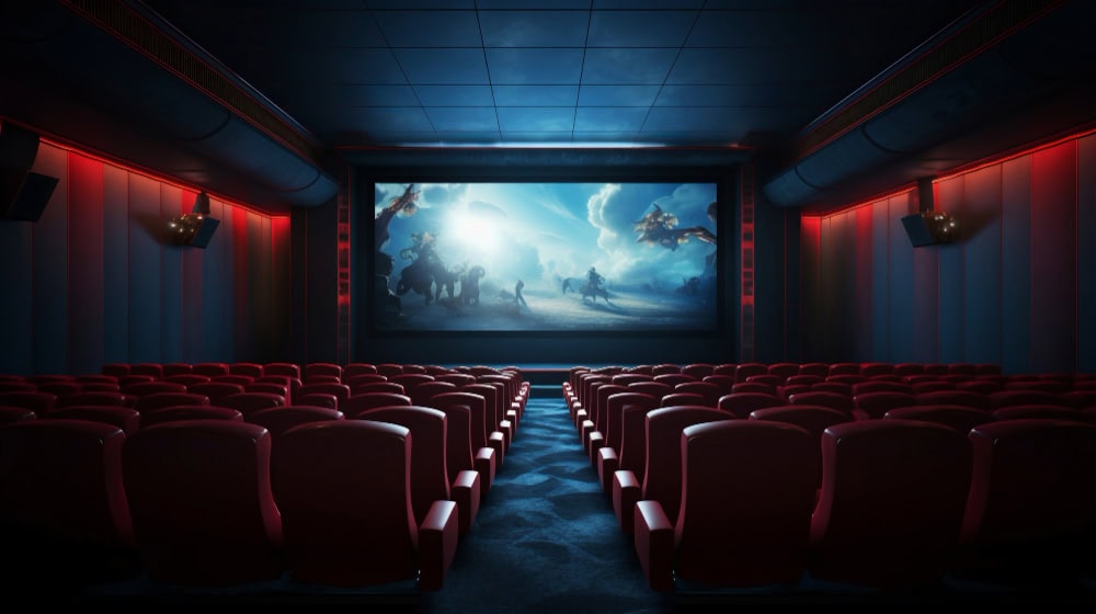 Dark movie theater with red seats facing a large screen displaying a blue-tinted prehistoric battle scene.