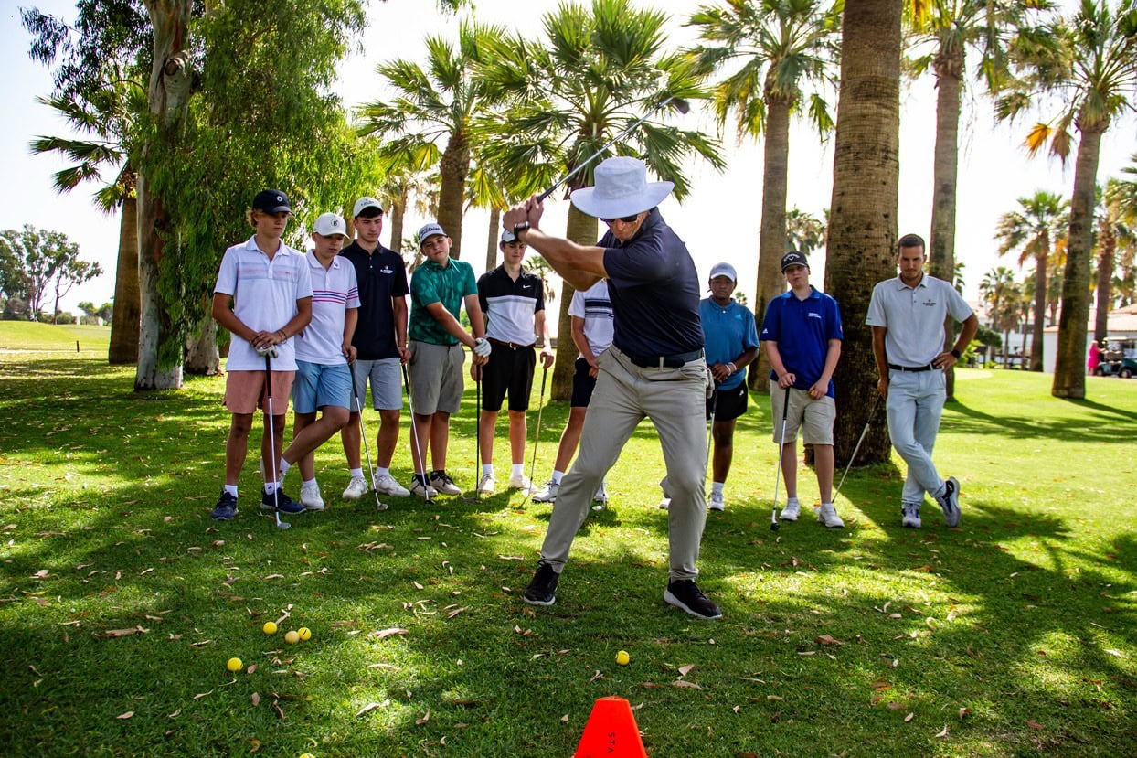 Golf instructor demonstrates swing to group of young players on a sunny course with palm trees behind them