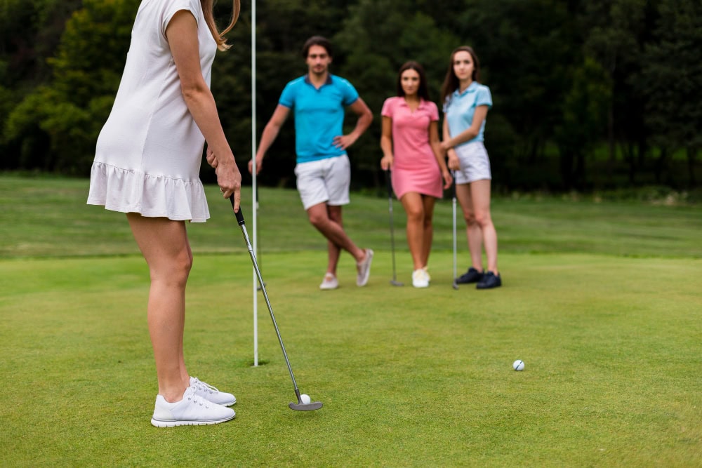 Woman in a white dress prepares to putt on a golf green while three players watch from behind the hole.