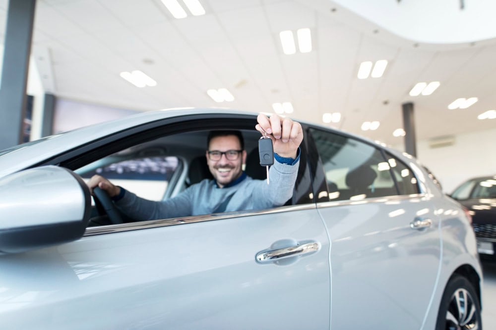 Smiling man sits in a silver car inside a bright showroom, holding up a car key to the camera or viewer.