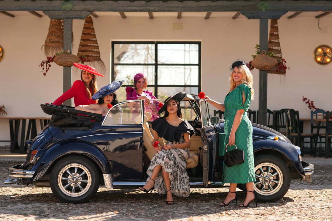 Five women in vintage dresses and hats pose with a dark blue convertible in a sunny courtyard.