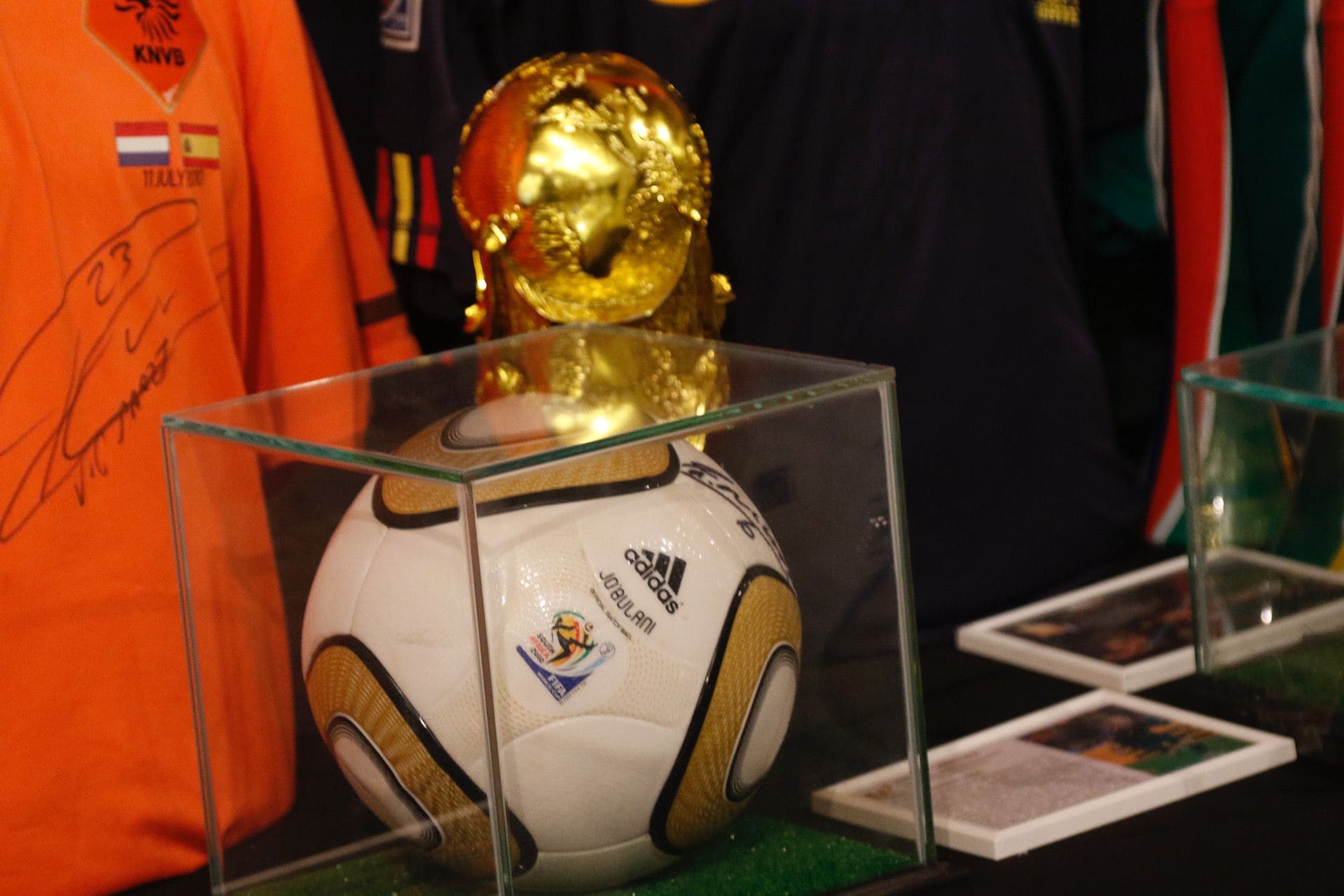 Soccer ball in a glass display case with a gold trophy in the background and an orange KNVB jersey beside it.