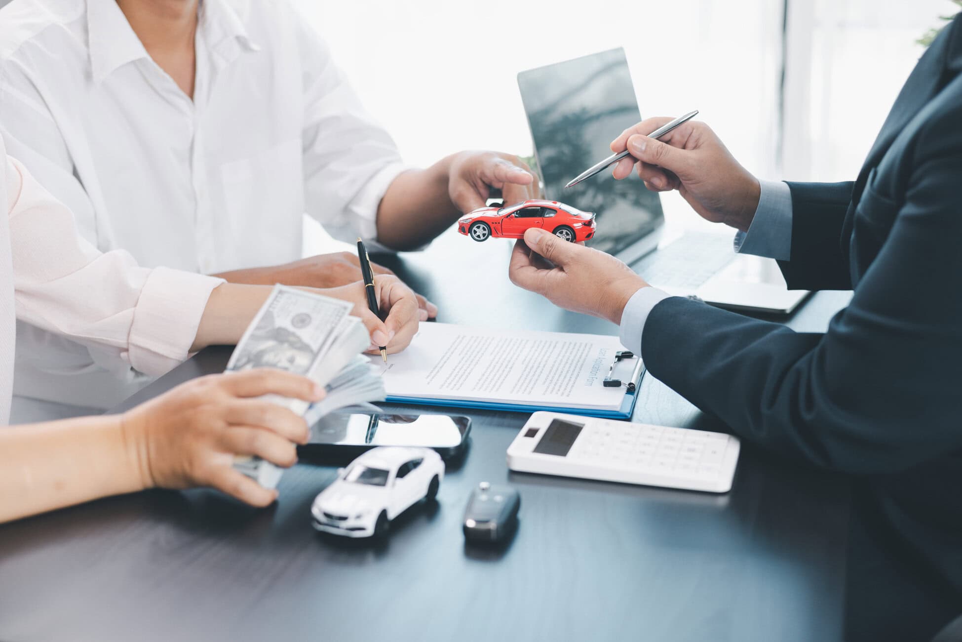 Business meeting: a person in a suit hands a red toy car to another person while reviewing documents on a desk with model cars and a calculator.