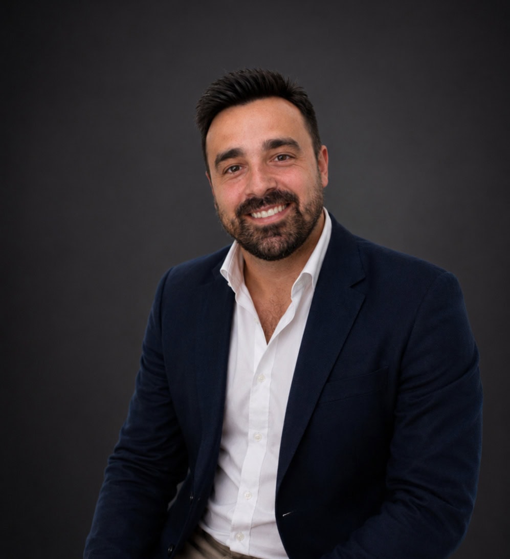 Professional headshot of a smiling man in a navy blazer and white shirt against a dark background, seated slightly angled.