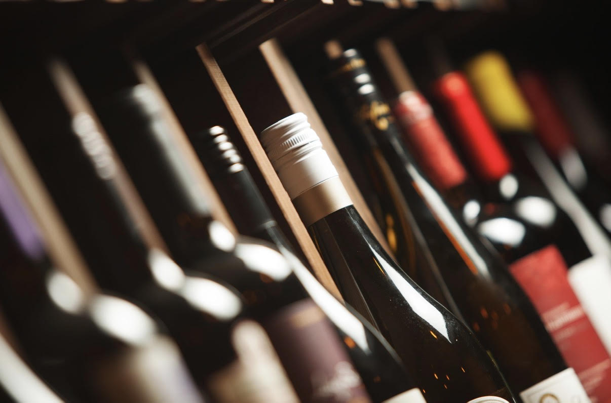 Row of wine bottles stored on a wooden rack, close-up with a white-capped bottle in the foreground.
