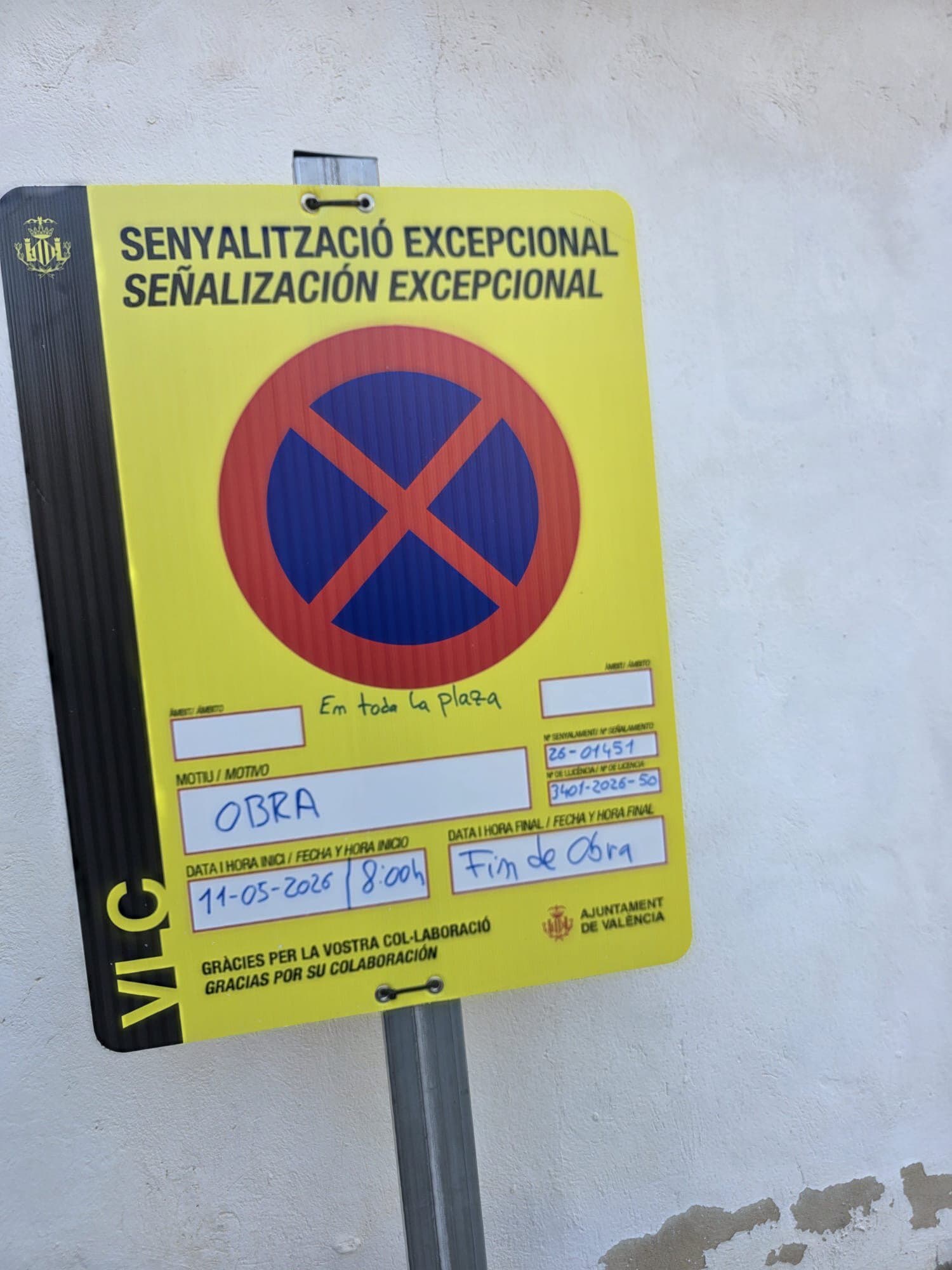 Yellow no-parking sign with a red circle blue X, text 'Senyalització Excepcional / Señalización Excepcional' on a metal post by a wall.