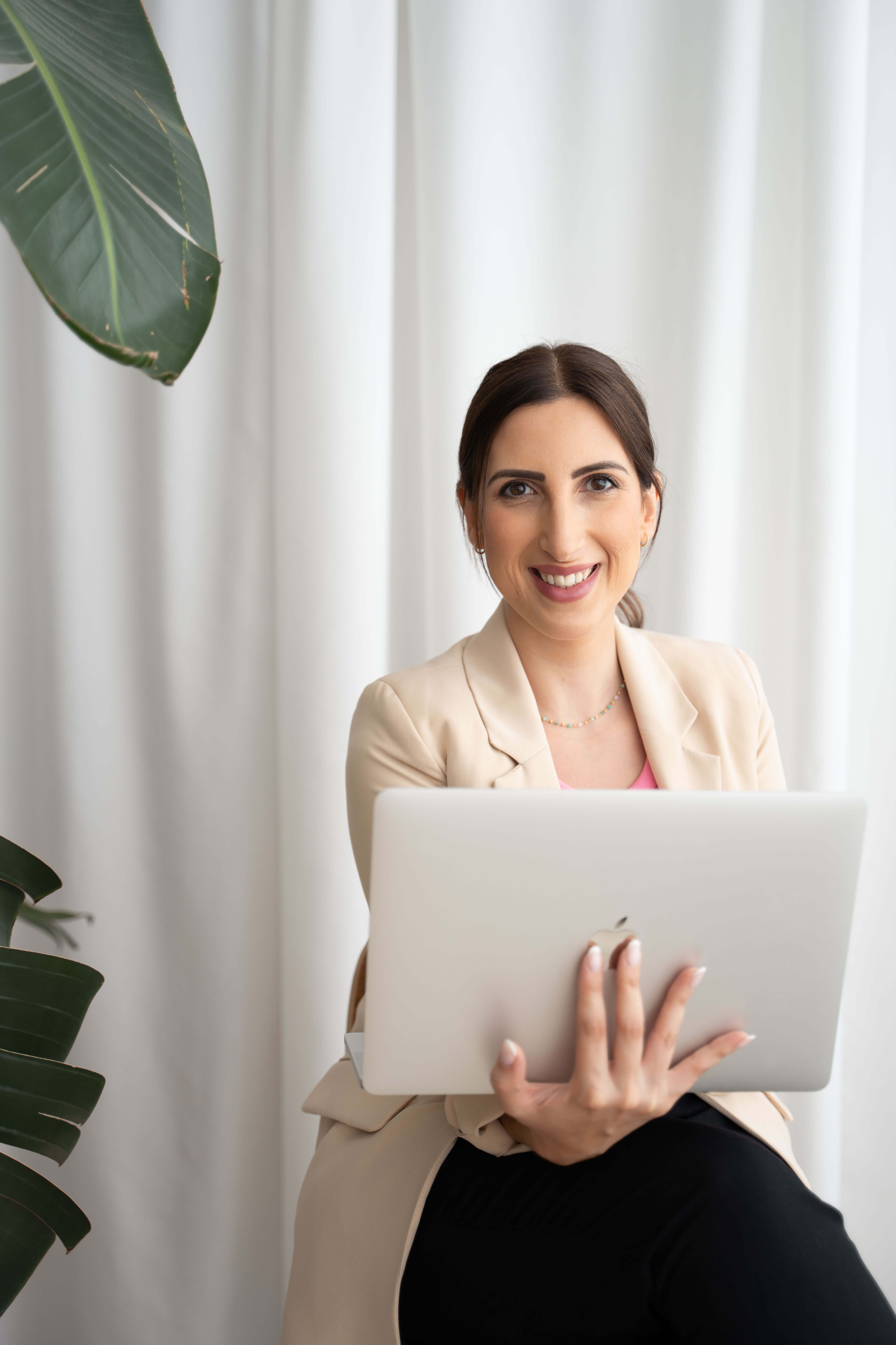 Smiling woman in a beige blazer holds a tablet indoors, with white curtains behind her and a plant nearby.