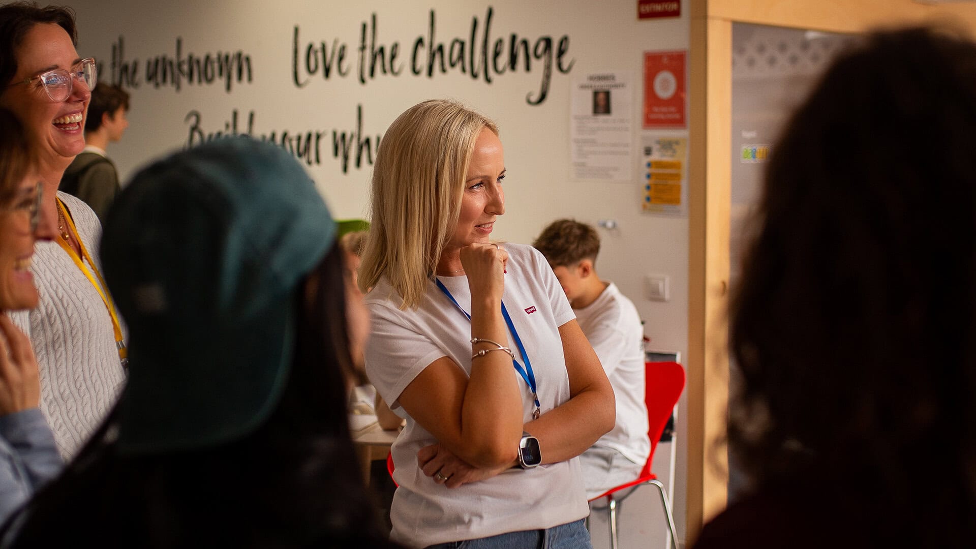 Blonde woman in a white shirt with a blue lanyard stands with her hand on her chin, listening at a workshop.