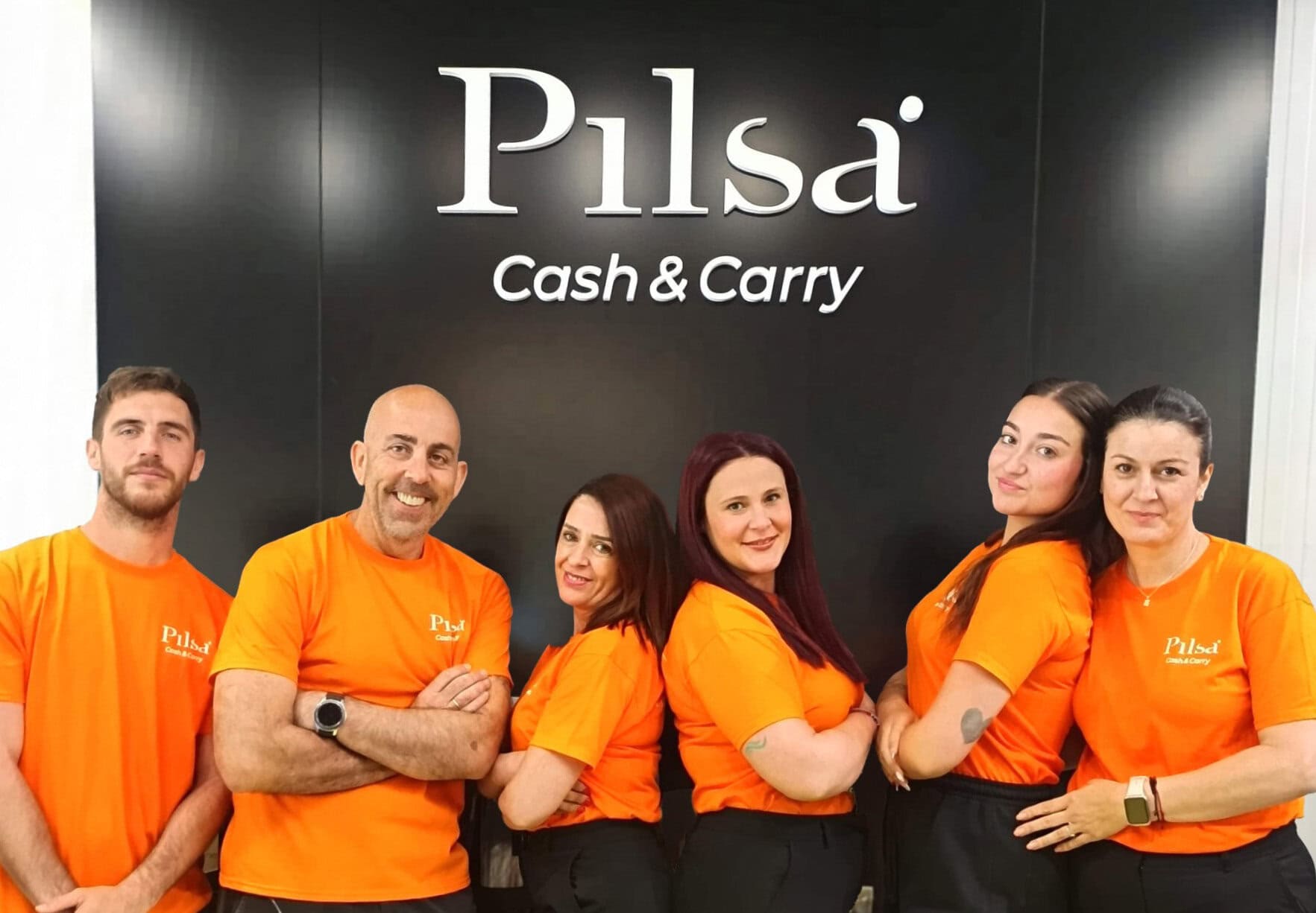 Six staff members in bright orange Pilsä Cash & Carry shirts stand side by side, arms crossed, smiling in front of a dark wall with the brand logo.