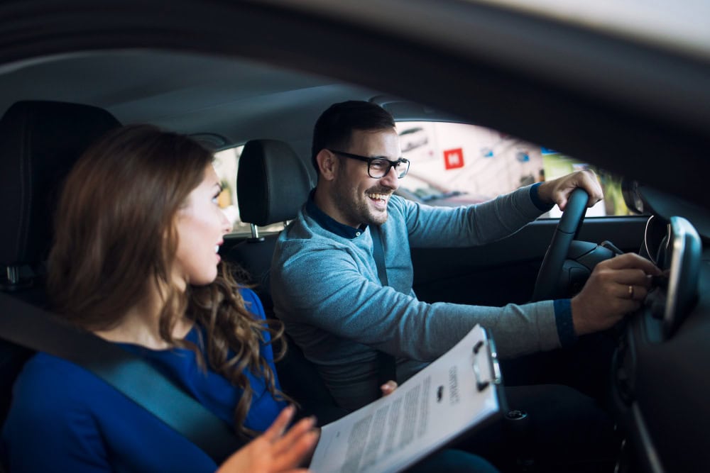 Man driving a car with a smiling woman passenger holding a clipboard beside him