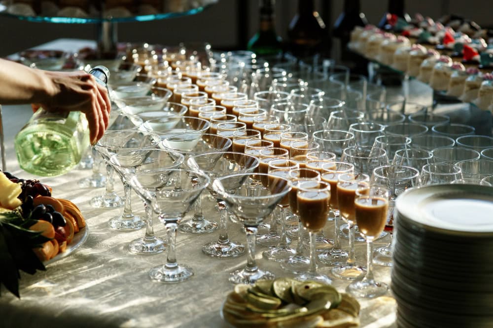 Catered table with rows of champagne flutes being poured, fruit platters visible to the side.