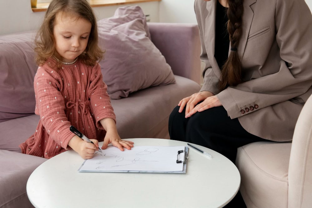 Young girl in a pink dress draws on a clipboard at a white round table while a woman in a blazer watches nearby on a pink sofa.