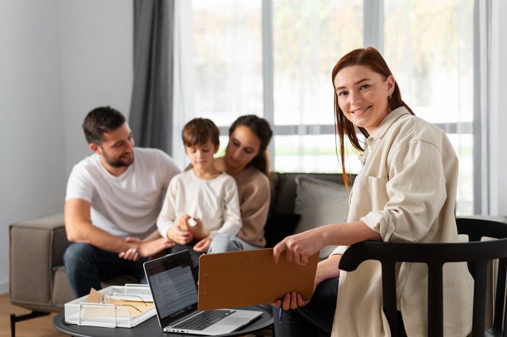 Therapist with clipboard and laptop sits at a table as a family of three talks on a sofa in a living room.