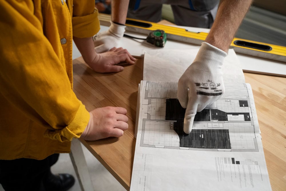 Person in a glove points to a floor plan on a worktable during construction.