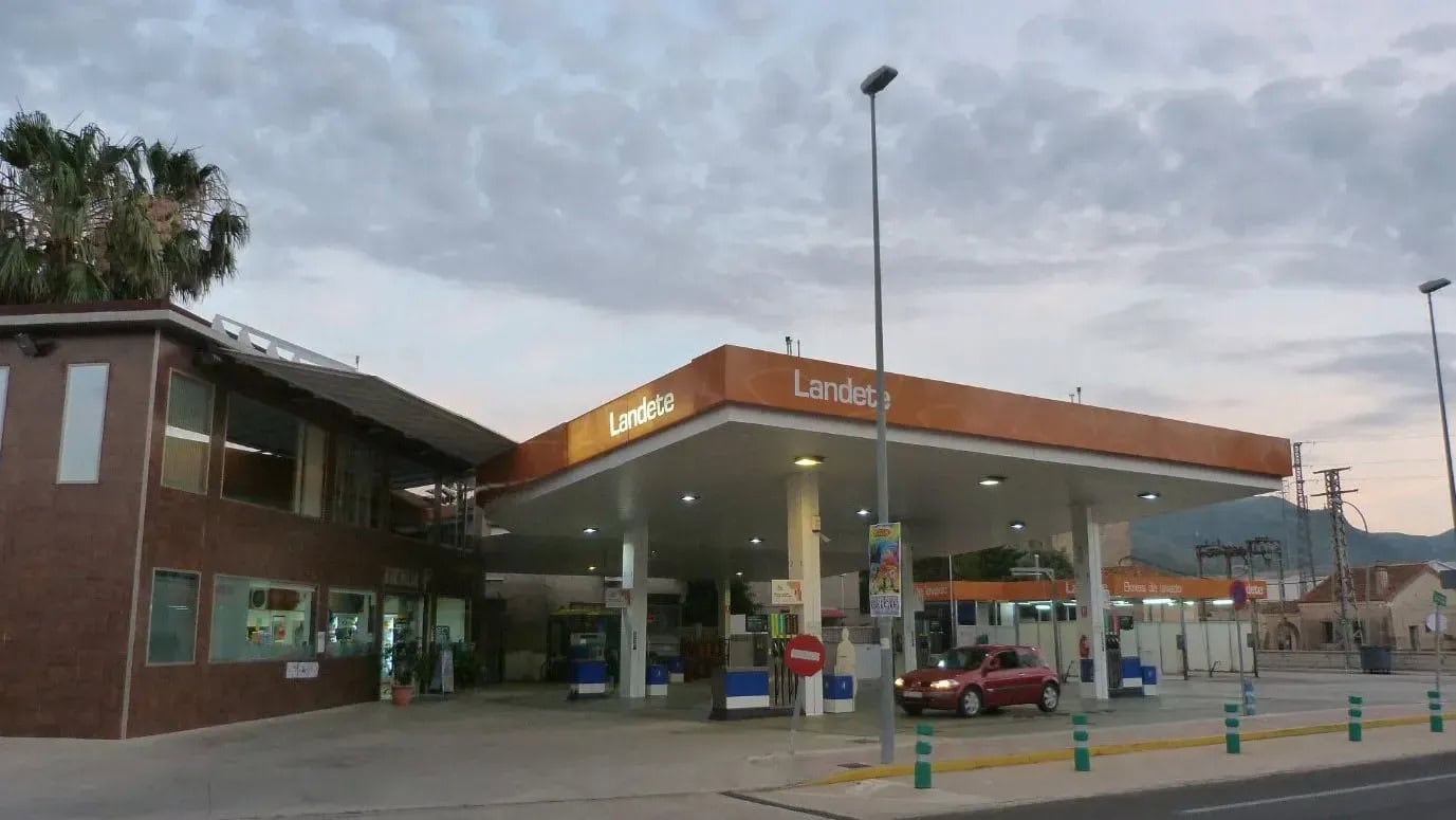 Gas station with orange canopy reading 'Landete' and a red car at a pump under a cloudy sky, left building visible.
