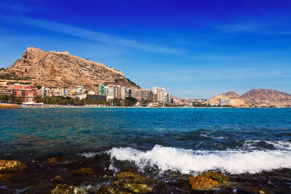 Coastal city with a hilltop fortress overlooking a sandy beach and calm blue sea, buildings along the shore.
