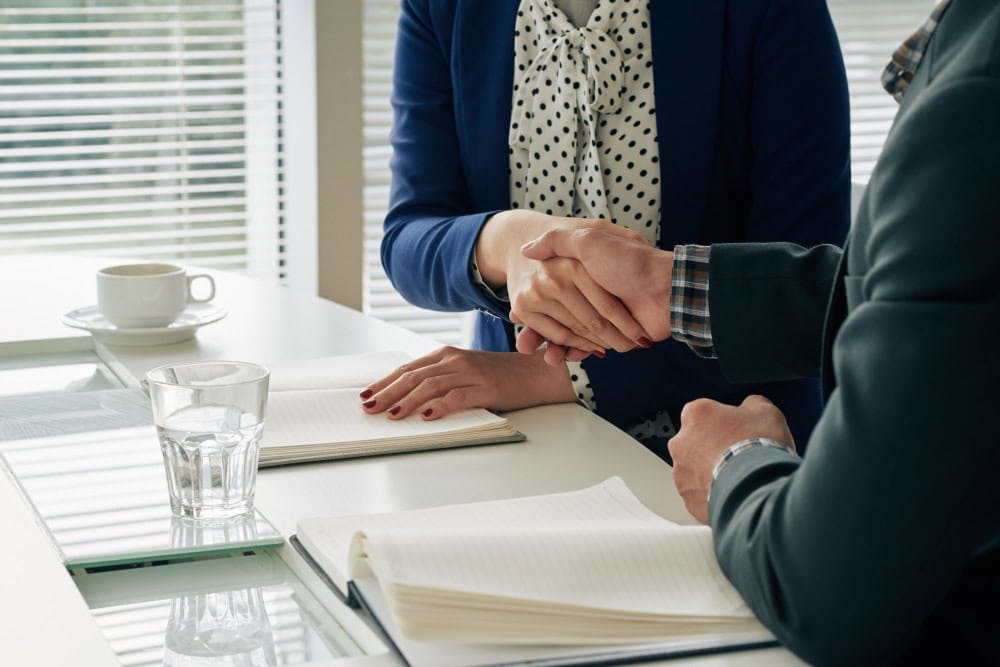 Two professionals shake hands over a white desk, with notebooks, a glass of water, and a cup nearby, signaling a business agreement.