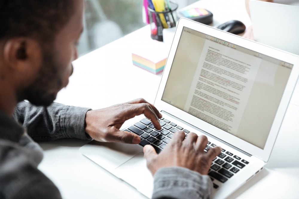 Person typing on a silver laptop at a white desk with a document visible on the screen.