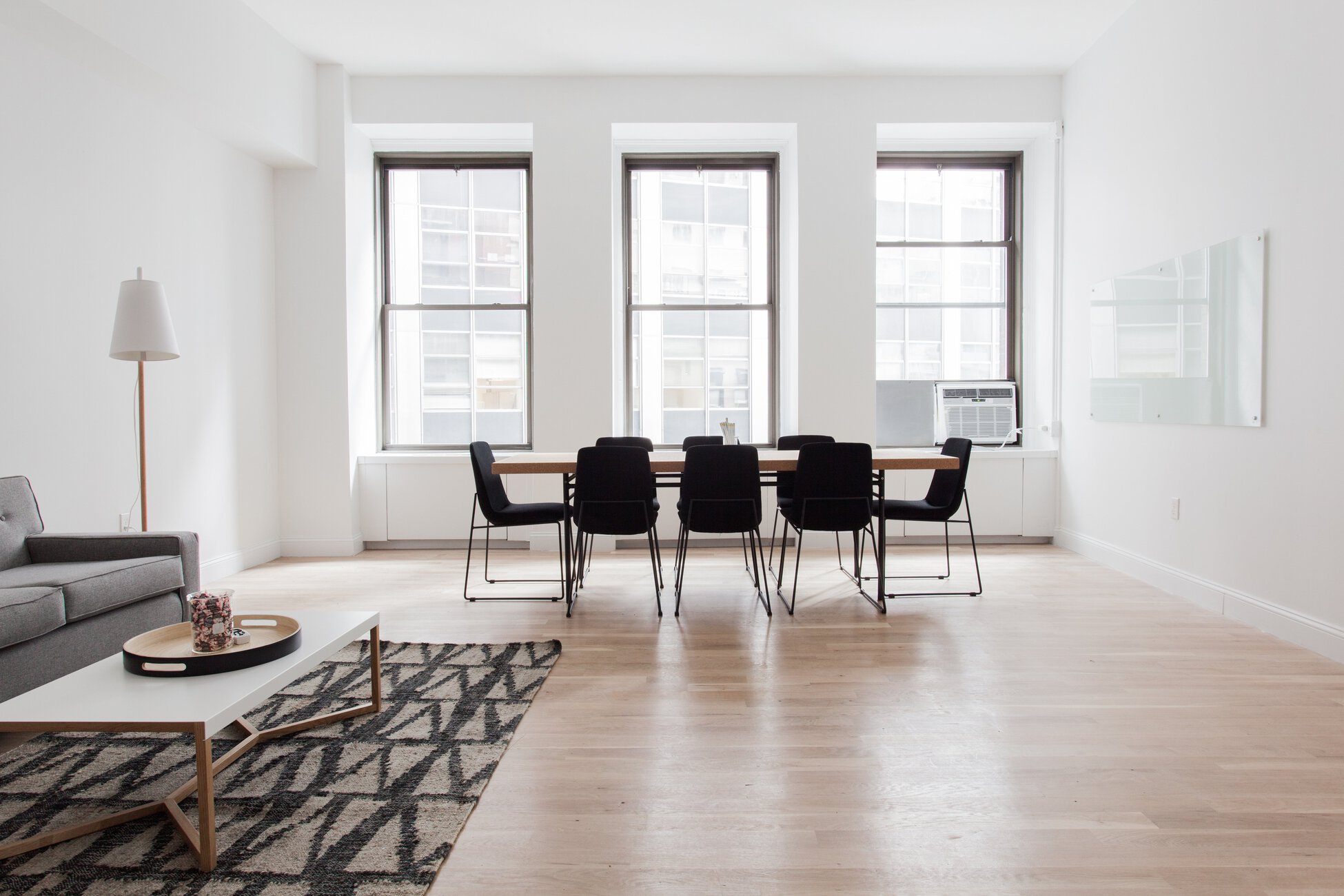 Bright open-plan living and dining space with a long table and eight black chairs, large windows, and white walls on all sides.