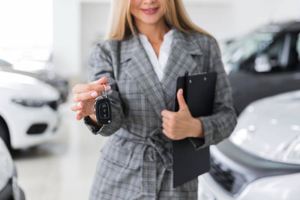 Woman in a gray plaid blazer extends a car key fob toward the camera in a showroom of parked cars.