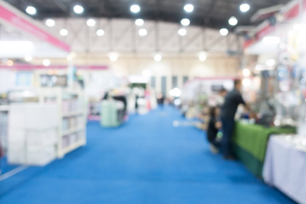 Blurred view of a trade show with booths along a blue-carpeted aisle and people browsing.