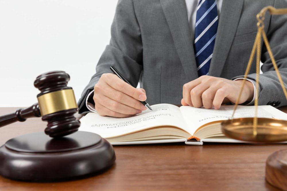 Close-up of a person in a suit writing in an open law book, with a gavel and scales of justice on the desk