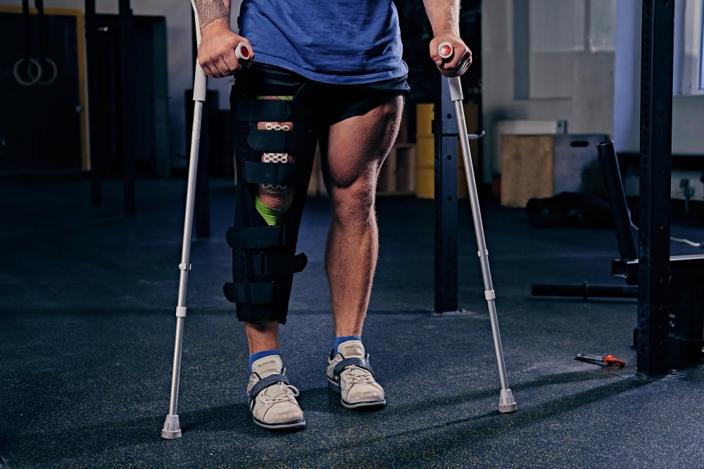 Person using crutches with a multi-strap knee-to-ankle brace on the left leg in a gym setting