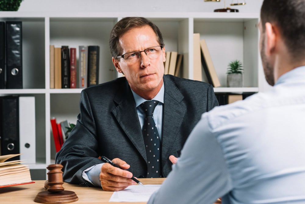 Lawyer in a dark suit and glasses sits at a desk with a gavel, discussing documents with a client across the table.