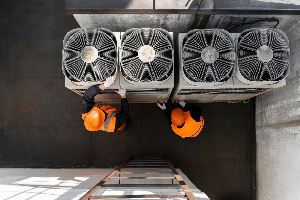 Two construction workers in orange safety vests and hard hats inspect four outdoor HVAC units on a roof (top-down view)./nLadder visible at the edge for context, sunlight cast shadows.