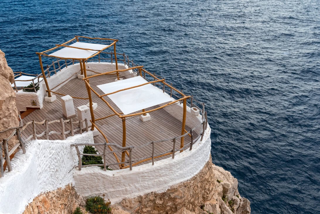 Cliffside terrace with white walls, wooden railings, and shade sails overlooking deep blue sea.