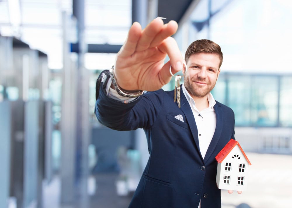 Man in a navy suit offers a set of house keys, with a small white model home in a bright office.