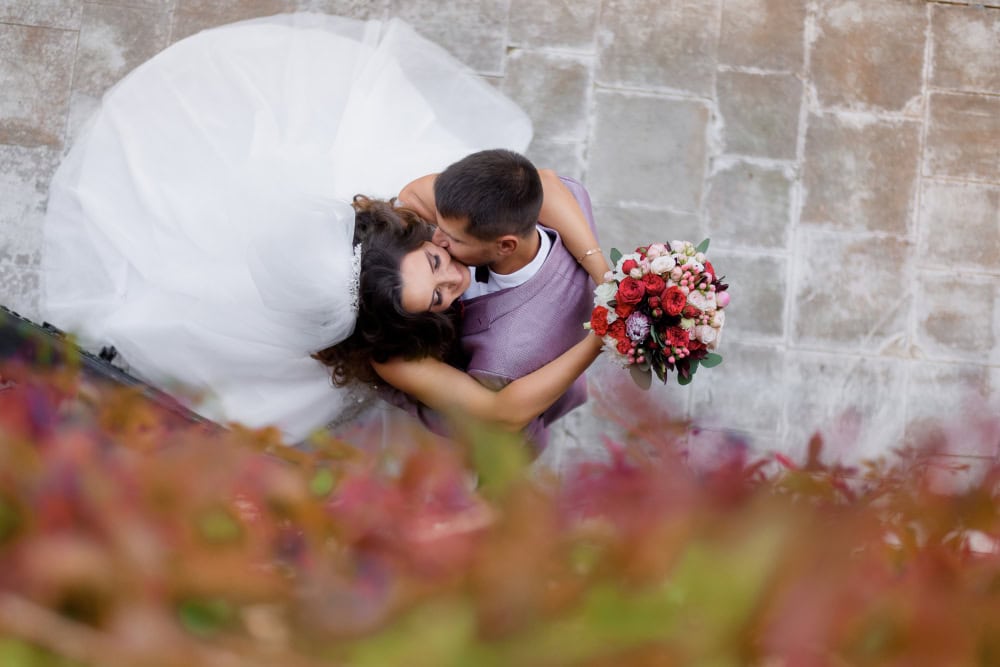Top-down view of a bride in a white gown kissing a groom in a lavender suit, bouquet in hand.