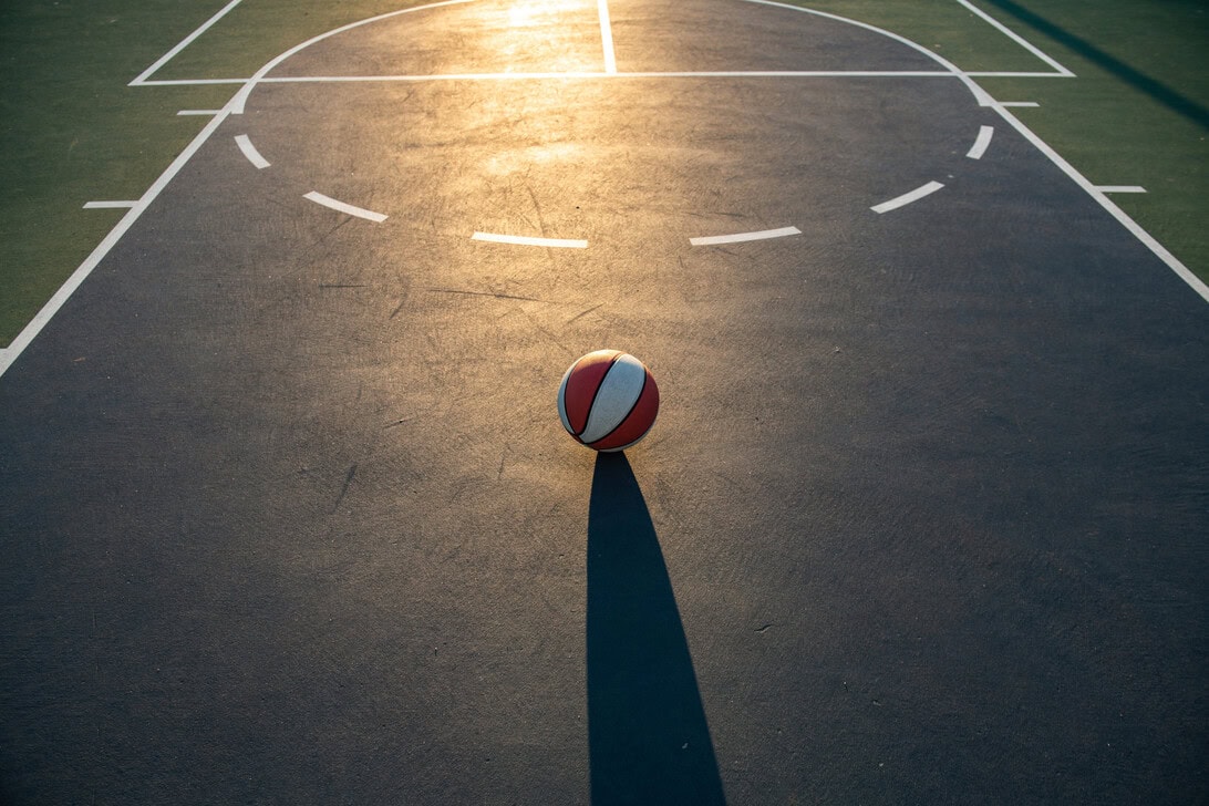 Basketball sits in the middle of an outdoor court, with a long shadow stretching toward the foreground at sunset.