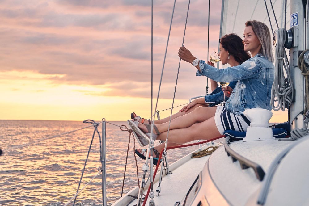 Three women sit on the edge of a sailboat at sunset, sipping wine and enjoying the ocean view.