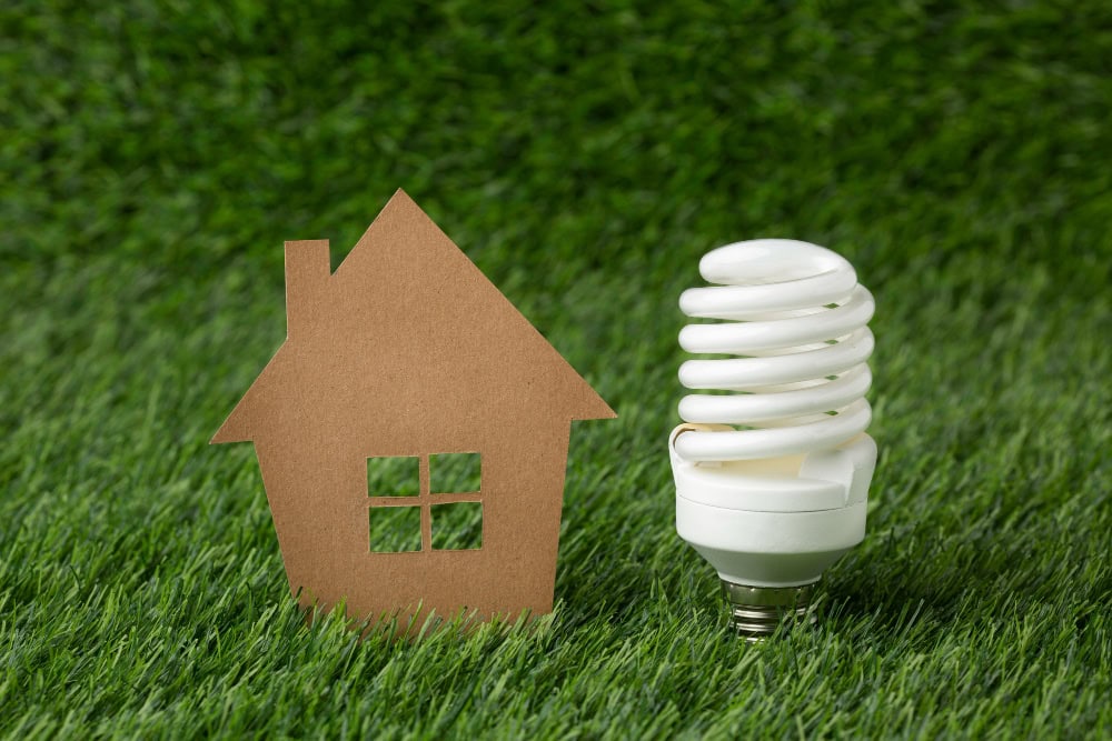 Eco-friendly home energy concept: paper house model beside a compact fluorescent lamp on grass