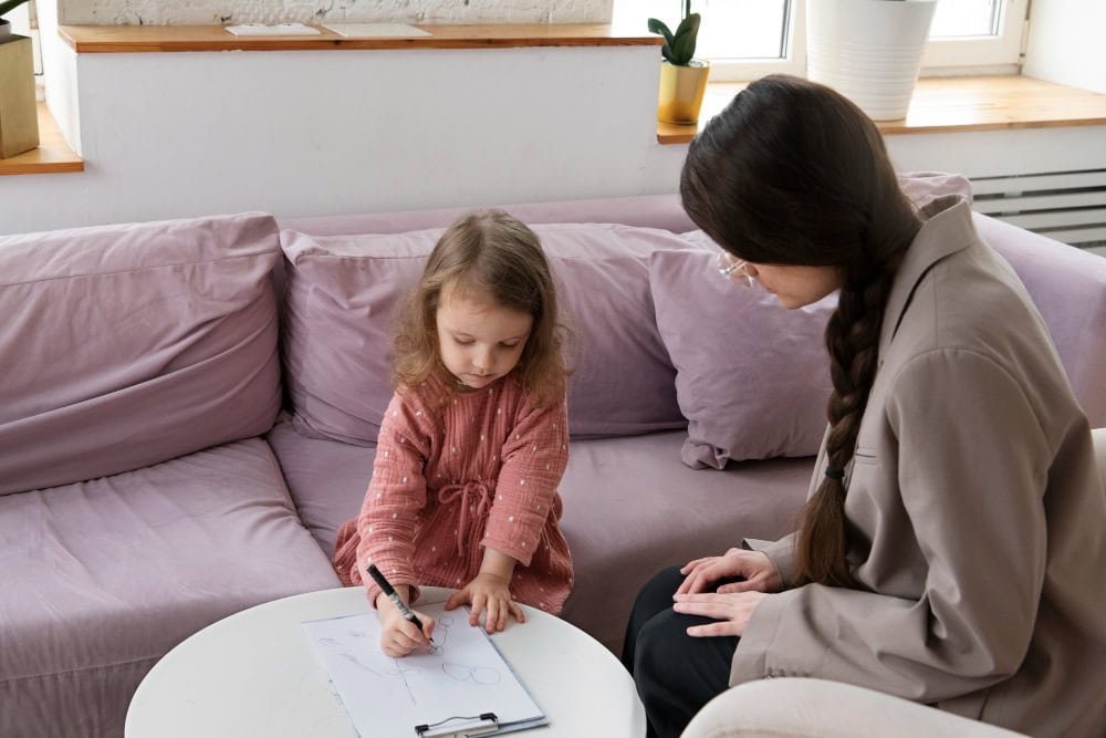 Young girl sits on a pink couch, drawing with a marker at a small white table while a woman watches nearby.