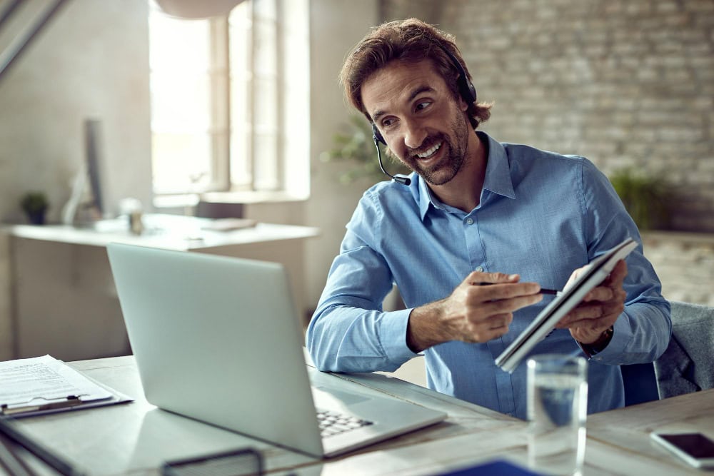 Man wearing a headset sits at a desk with a laptop, holding a tablet and pen, smiling as he talks or explains something.
