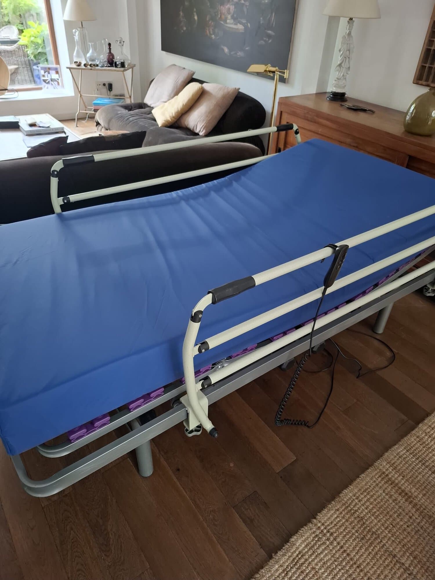 Adjustable hospital bed with blue mattress in a living room, white rails, and control cords visible by the side table and sofa.