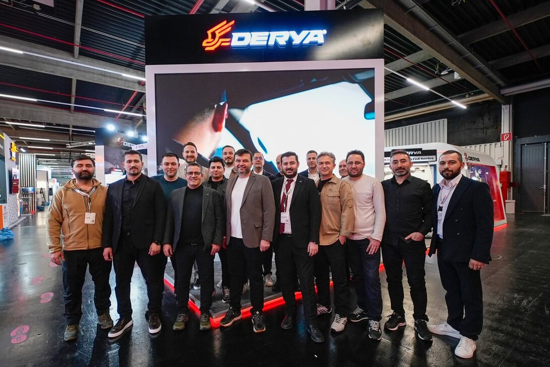 Group of professionals posing in front of a large screen at a DERYA trade show booth, logo visible above them.