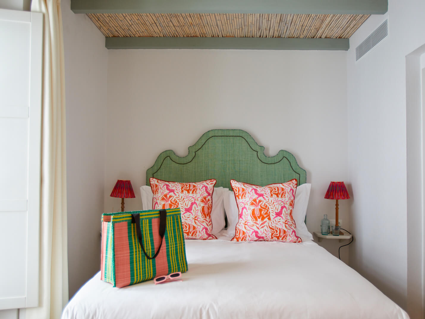 Bedroom with a mint-green upholstered headboard, white bedding, and two pink/orange tropical-pattern cushions; a colorful striped tote and pink sunglasses sit on the duvet, with red bedside lamps on either side.