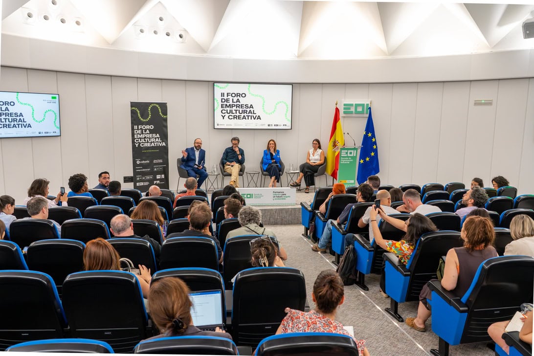 Panel discussion in a lecture hall with five speakers on stage, audience watching and taking notes, banners and flags nearby.