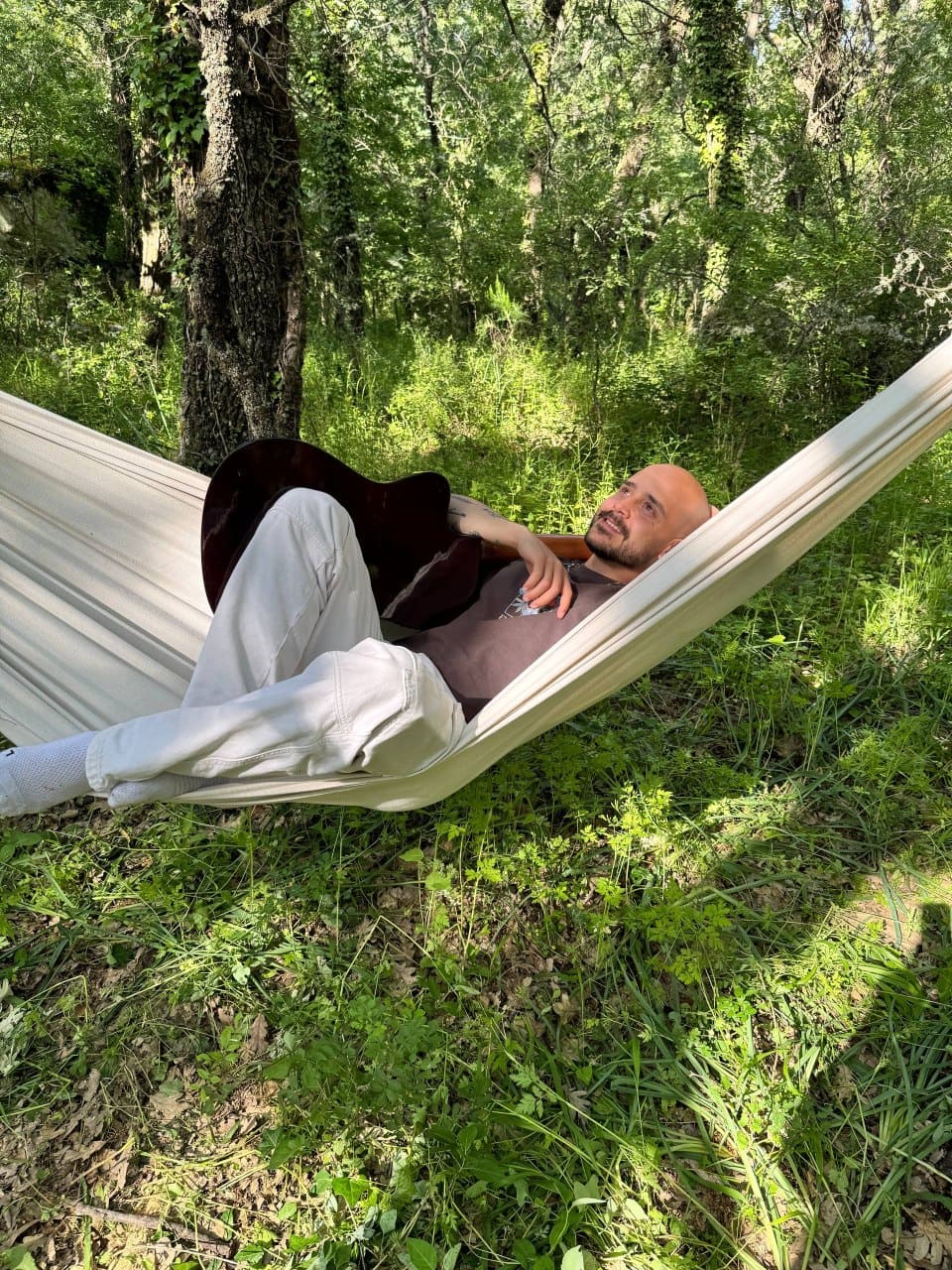 Man relaxing in a hammock tied between trees in a lush forest, holding an acoustic guitar across his chest.