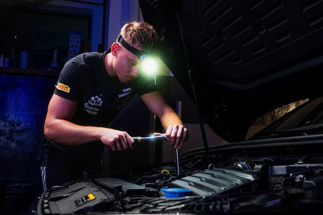Mechanic with a headlamp inspecting a car engine under the open hood, tool in hand