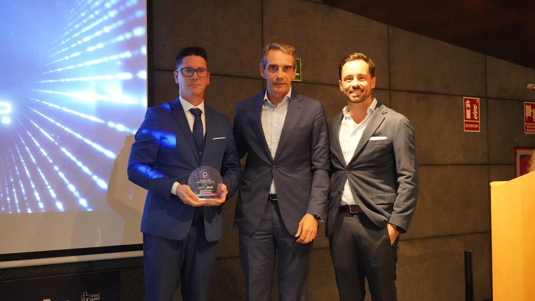 Three men in suits on a stage; the leftmost man holds a glass award.