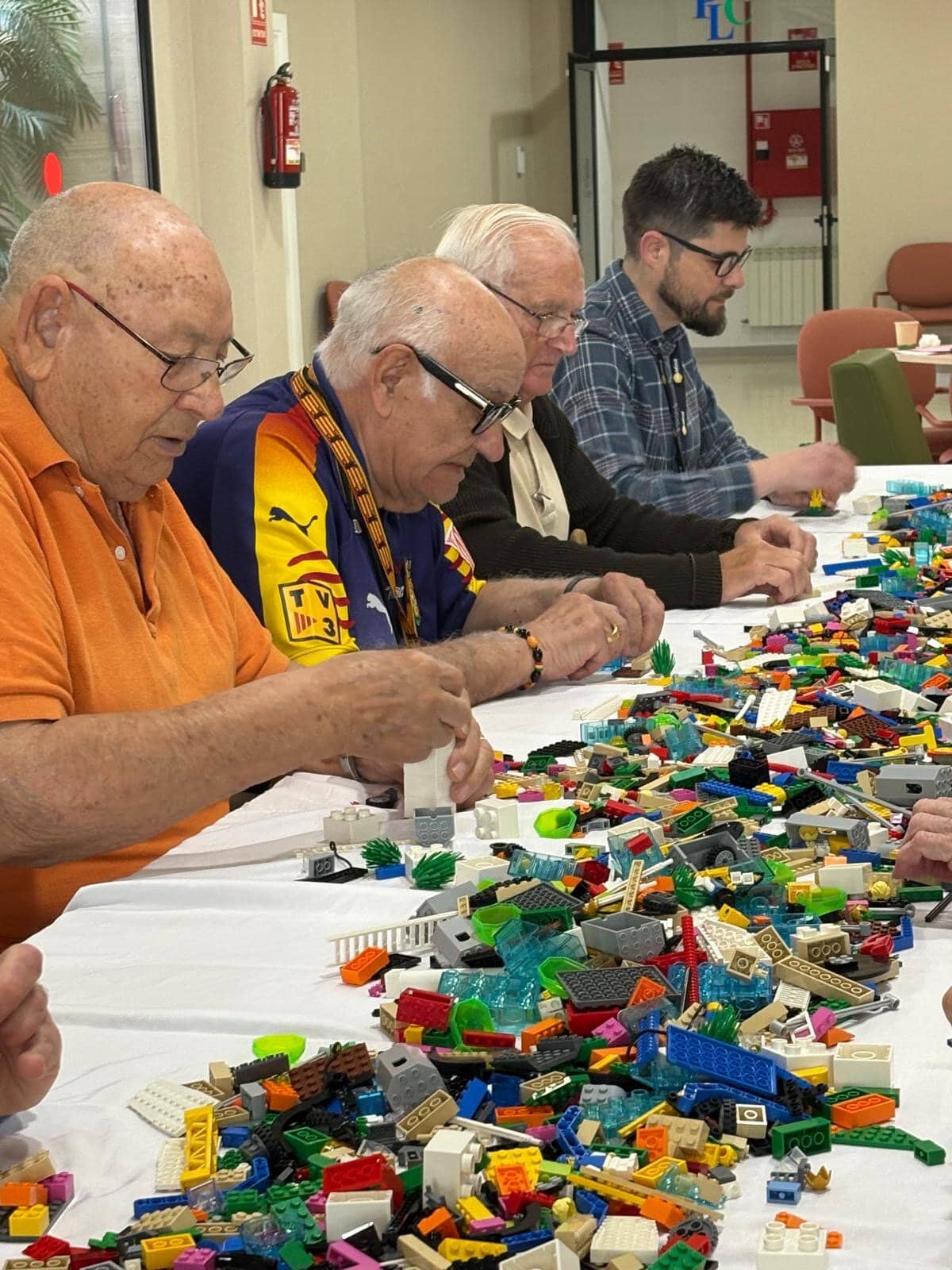 Four men of varying ages sit at a long table covered with colorful LEGO bricks, concentrating on building.
