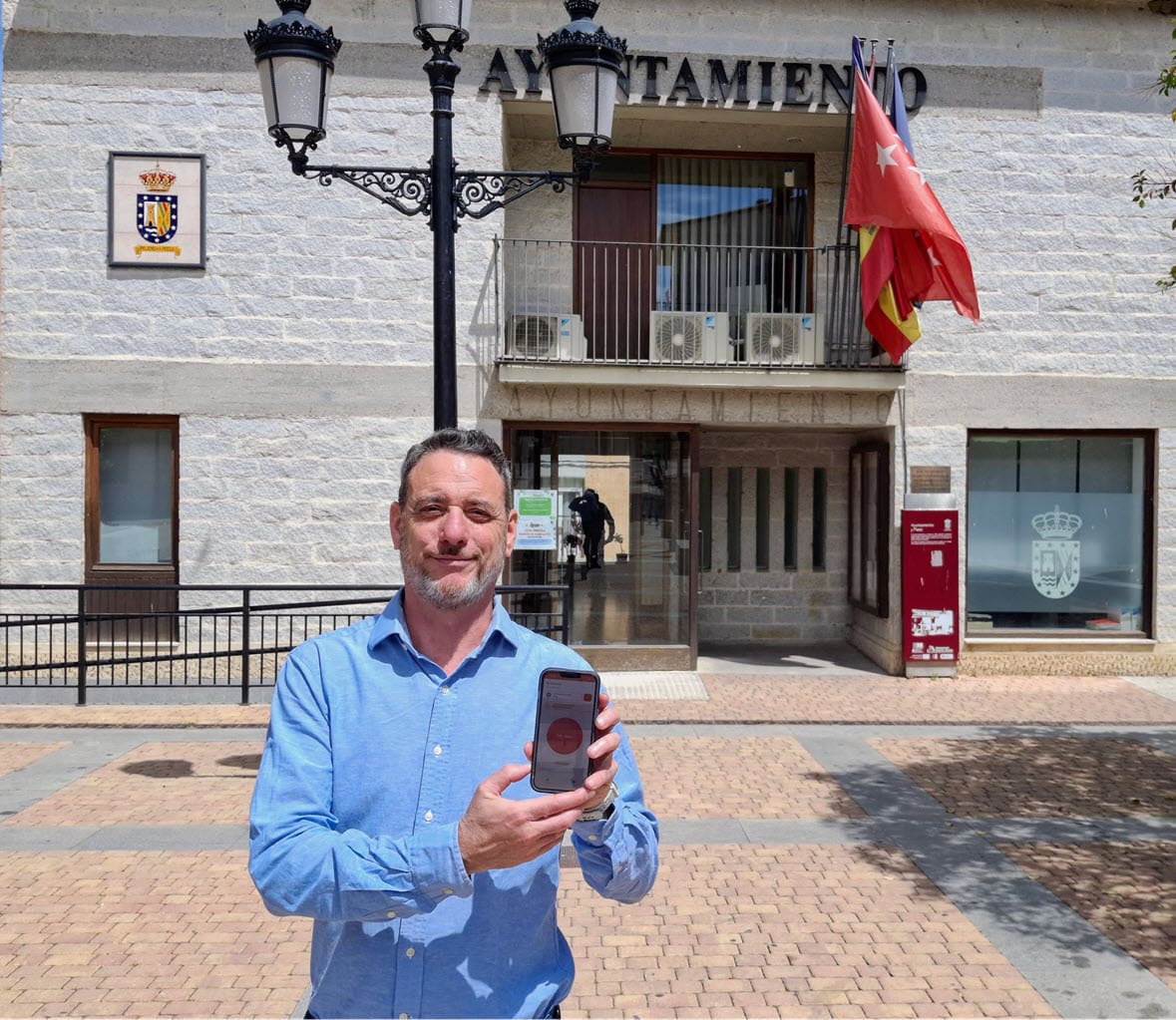 Man in a blue shirt stands outside a town hall (Ayuntamiento), holding up a smartphone; flags and a decorative street lamp are visible in the scene.